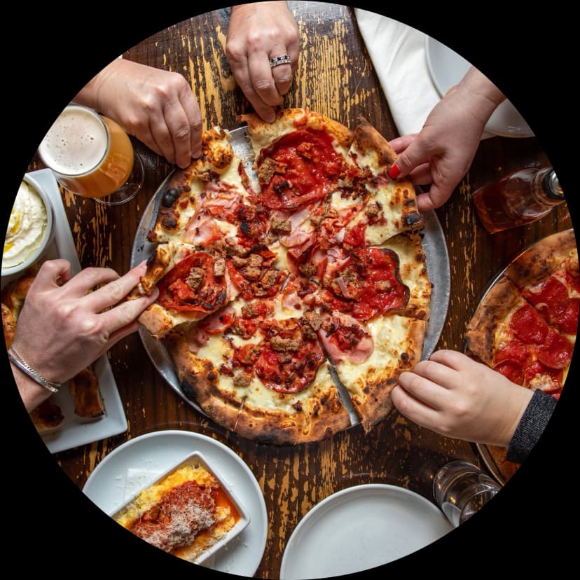 Pizza on a table with people reaching for a slice.